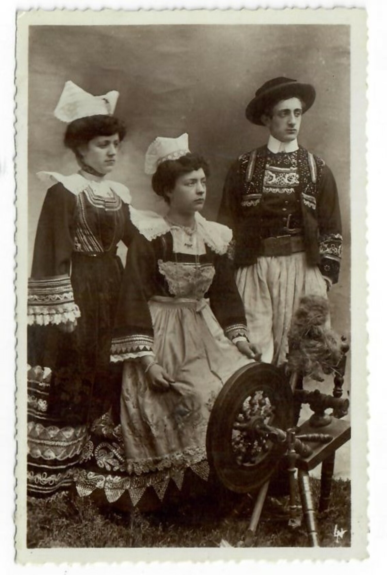 Postcard from early 20th century France, depicting two women and one man in local dress. One of the women is sitting in front of a horizontal spinning wheel. There is a text on the reverse that states: "3734 - Costumes de PONT-AVEN" (TRC 2020.4246).