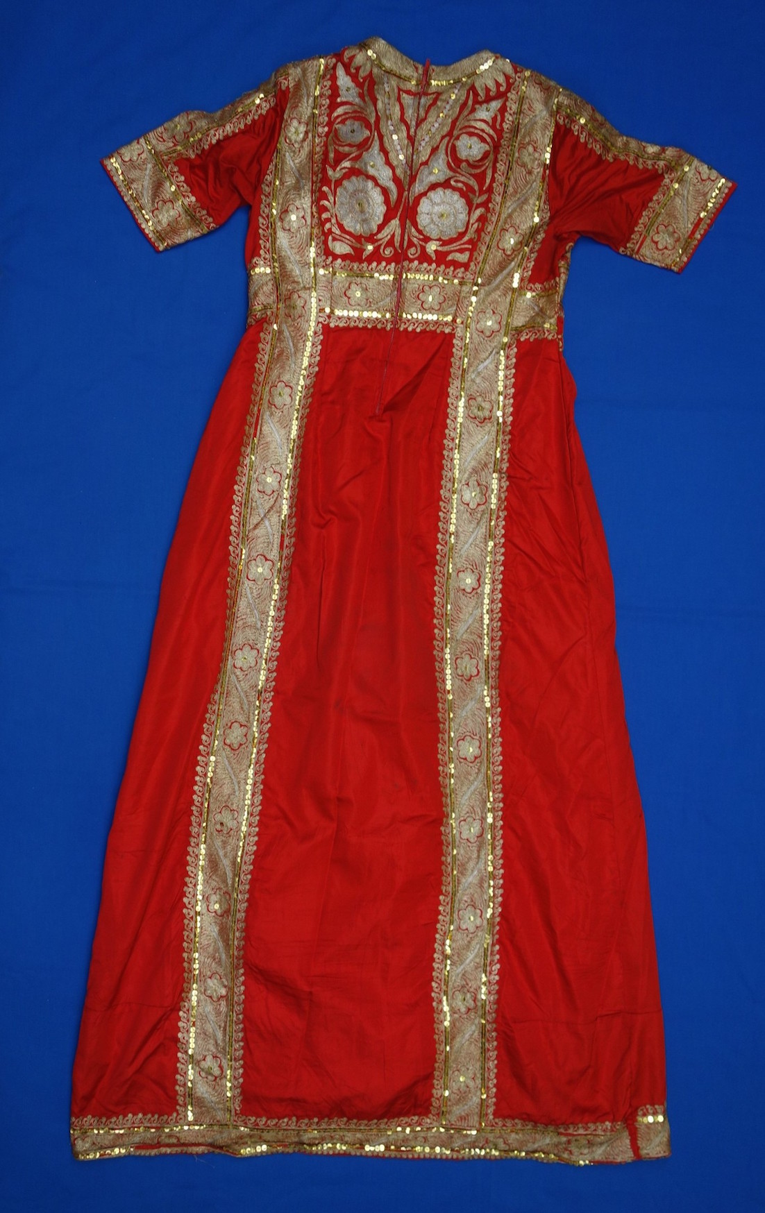 Woman's dress from Saudi Arabia. TRC 2005.0077 3. Modern fustan made from red Indian silk heavily decorated with machine embroidery in gold and silver threads and gold sequins. The garment is decorated with long borders of flowers, palm leaves, scrolls. It has a central panel with palm trees, Arabian daggers, traditional pine and leaf patterns.